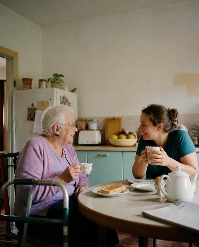 An older woman with a walking frame sharing tea and toast with a support worker at a round kitchen table, both smiling