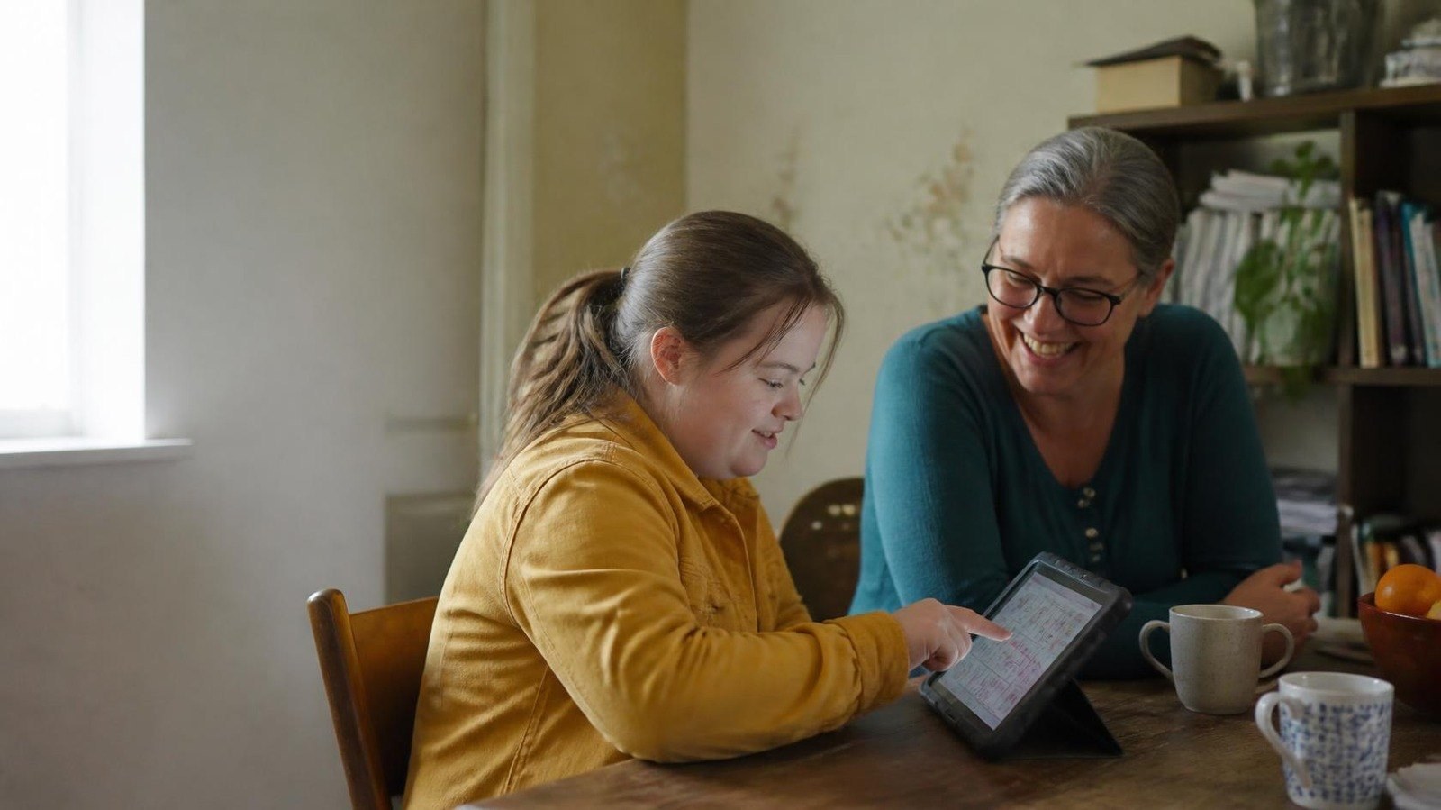 A young woman pointing to symbols on a tablet communication app at a kitchen table while an older woman beside her laughs